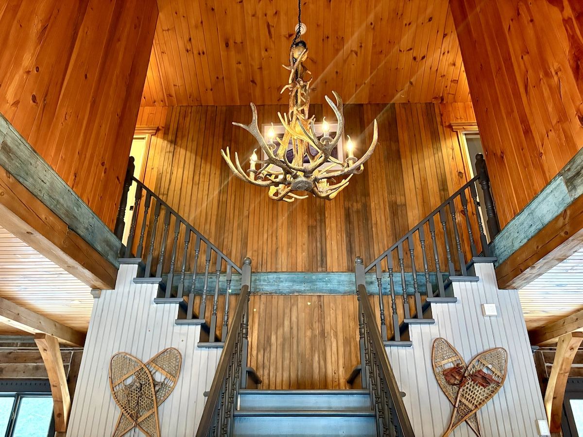 The great room's cathedral ceiling with an antler chandelier, mezzanine railing, and hand-cut beams