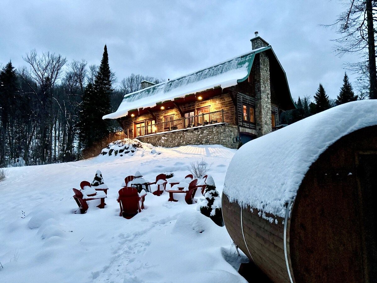 The house in snow with red Muskoka chairs around the fire area and the barrel sauna
